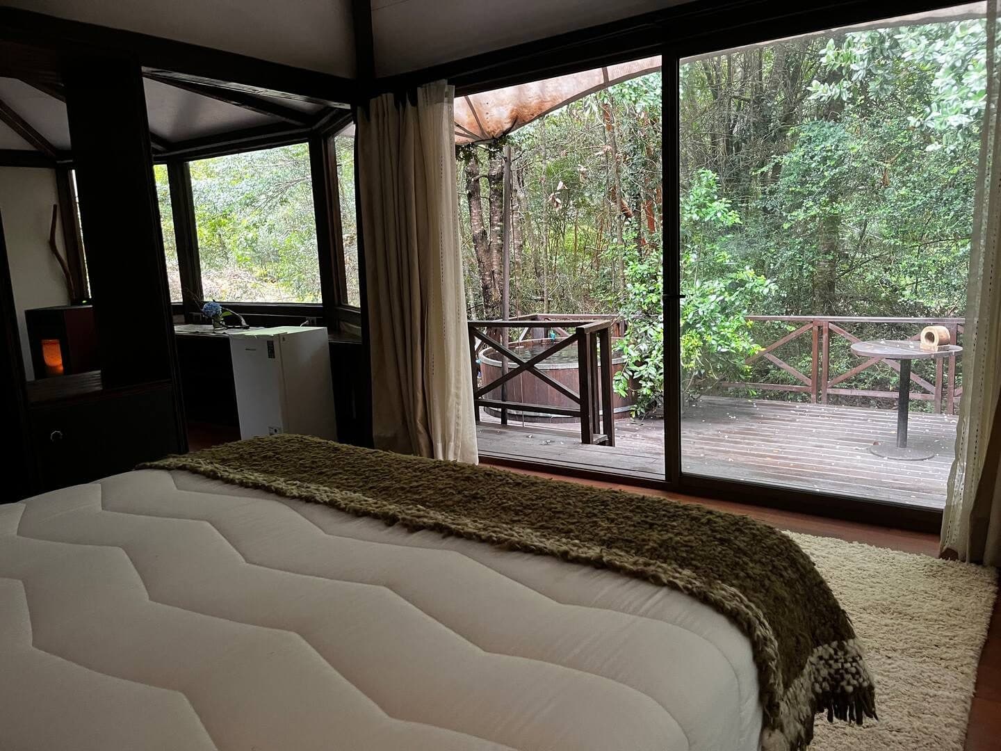 Cama frente al ventanal abierto hacia la terraza, con la tinaja y el bosque de fondo