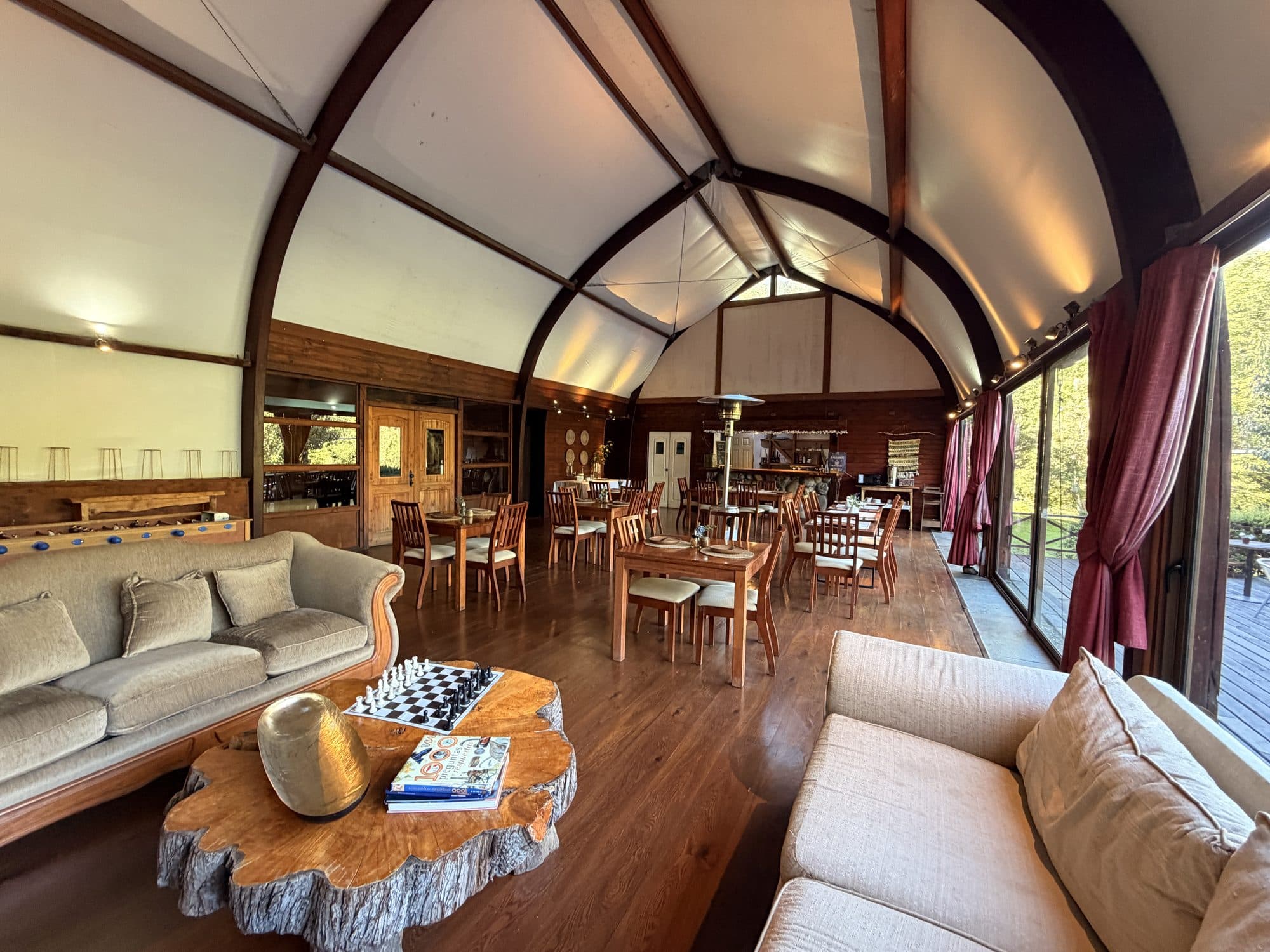 Salón principal del lodge con techo abovedado de madera, sofás, mesas de comedor y una mesa de ajedrez sobre un tronco